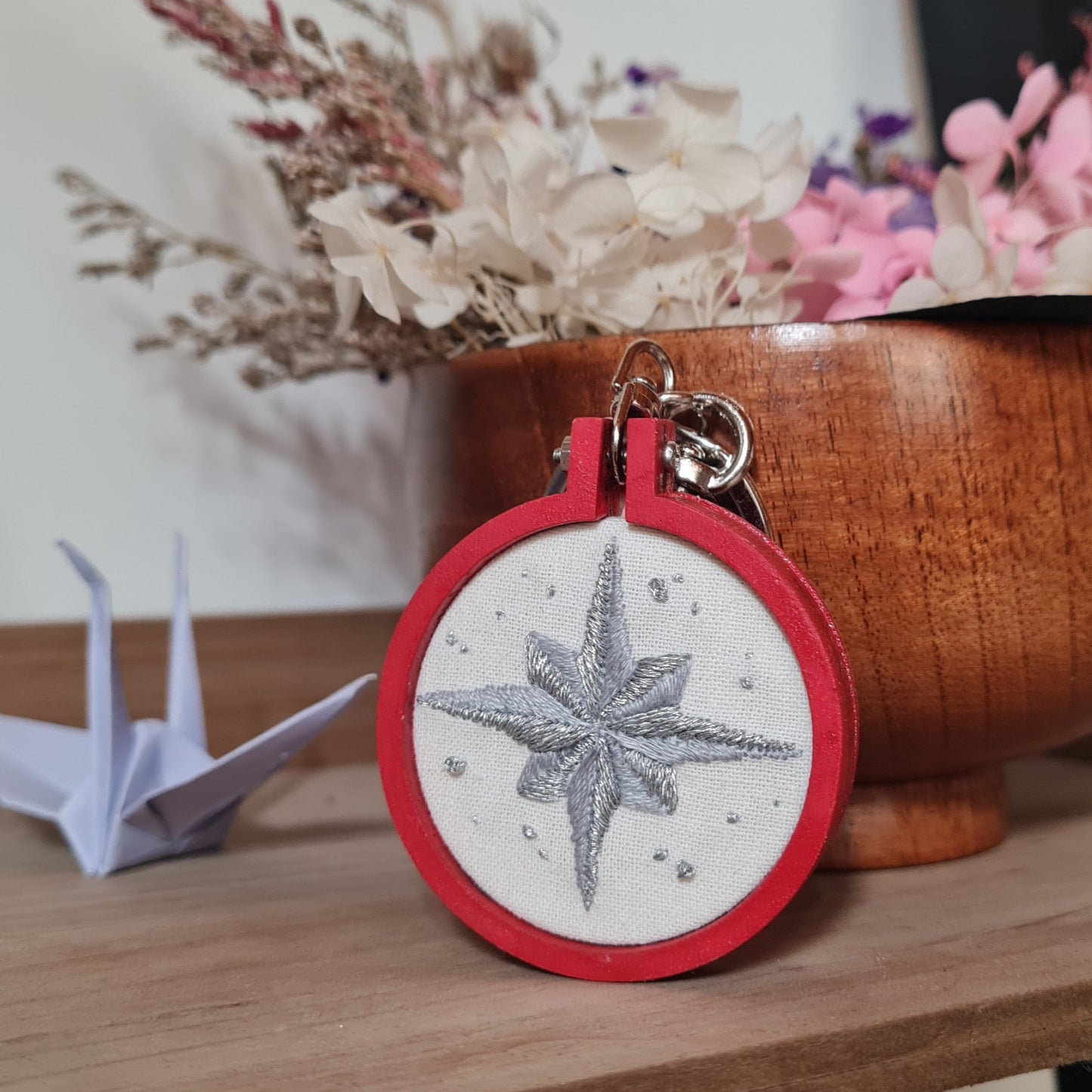 Porte-clés Broken Compass, inspiré de Stray Kids. Il met en avant la boussole symbolique présente sur le lighstick des STAYs, élément clé de l'univers du groupe. La flèche de direction est brodée dans une alliance de gris neutre et de fil argenté, faisant ressortir les contrastes de la broderie. Le tissu est encadré par un mini tambour en bois, de couleur rouge. Chaque objet est peint, vernis, brodé et assemblé à la main, pour un rendu de haute qualité.