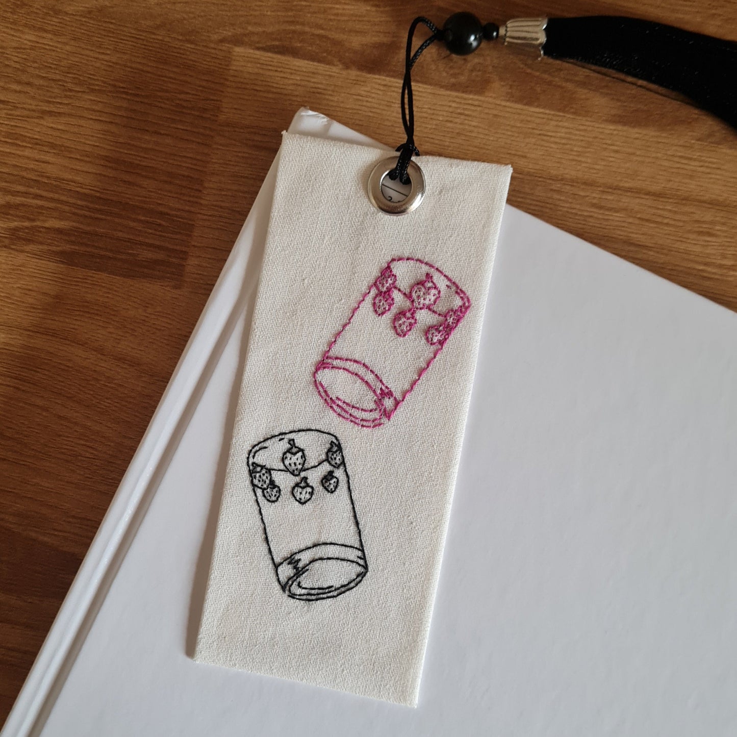 Marque-page brodé inspiré du manga Nana, représentant les verres fraises iconiques de Nana et Hachi. Un verre est noir et l'autre est rose.