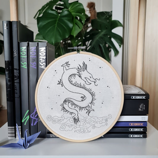 Broderie dragon et nuages sur toile en coton, inspirée de l'album 5-star de Stray Kids. Dessinée et brodée à la main, cette décoration délicate reprend des motifs issus de l'art asiatique. Elle mêle le noir et l'argent pour offrir une touche authentique et traditionnelle à votre intérieur. Elle est présentée sur un cercle à broder en bois clair, et posée contre des albums avec une couverture noire et une plante monstera en fond.